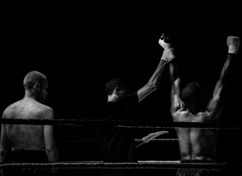 Black and white image of a boxer getting his hand raised in victory while another looks on.