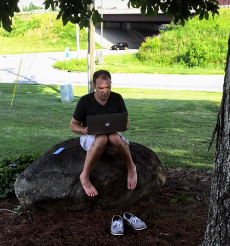 Person sitting barefoot on a rock under a tree working on a laptop