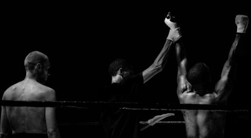 Black and white image of a boxer getting his hand raised in victory while another looks on.